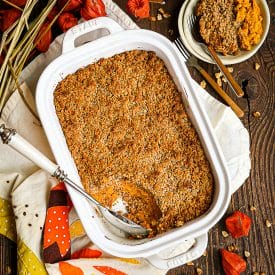 A scoop of Sweet Potato Soufflé removed from the baking dish with the serving spoon in the dish and flowers and nuts scattered around.