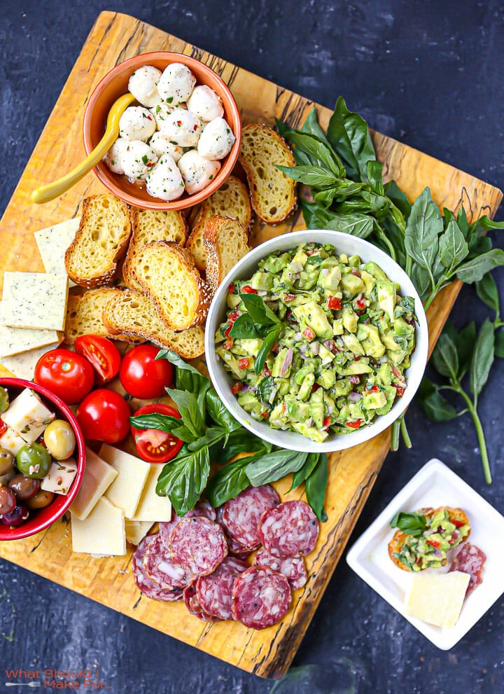 Italian Guacamole on a large wood board with bread, cheese, olives, basil, and salami and a serving on a small white plate.