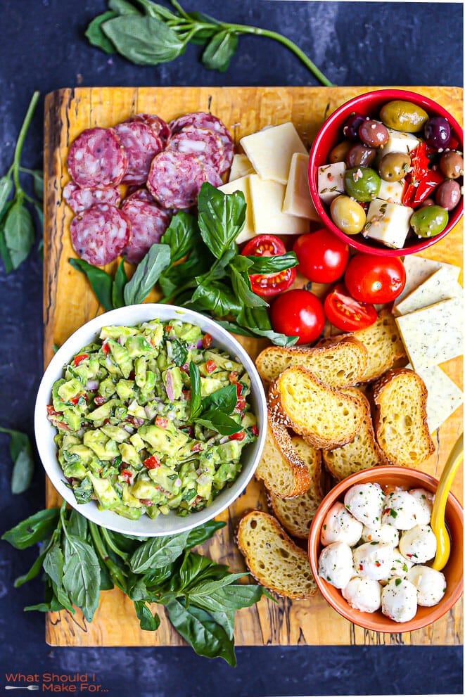 Italian Guacamole on a large wood board with bread, cheese, olives and salami.
